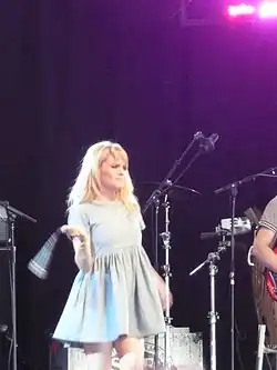Image of a young, blonde woman performing on stage. The woman is wearing a short, light blue dress. The performer is seen to be swinging a microphone.