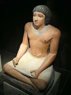A painted, realistic stone statue of a black-haired, perhaps middle-aged man sitting cross-legged while holding a stone-carved depiction of a papyrus reading scroll in his lap