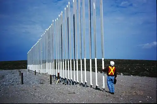 A worker in high visibility clothing and a hard hat examines a long line of pipes about four times his height sticking out of rocky ground.