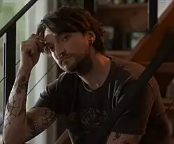 Erik (played by Richard Harmon) in a black shirt, resting his head on his right hand, looking into the camera