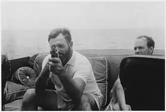 A bearded man sitting on cushions on a boat in the sea aims a gun at the camera; the man looking next to him looks exasperated.