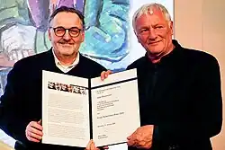 Two men standing in the foreground holding a booklet containing a certificate corresponding to the prize. The man on the left, Biebel, is wearing glasses, while the man on the right, Brustmann, has white hair.