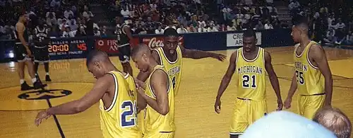 Five black men in gold athletic uniforms in the foreground on the sidelines of an athletic court while a few opposing athletes in green wait in the middle of the court.