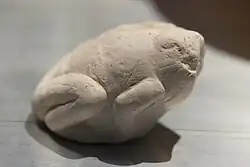 A light-grey stone shaped to resemble a crouching frog. The head is eroded, but the limbs are still visible tucked under the body.