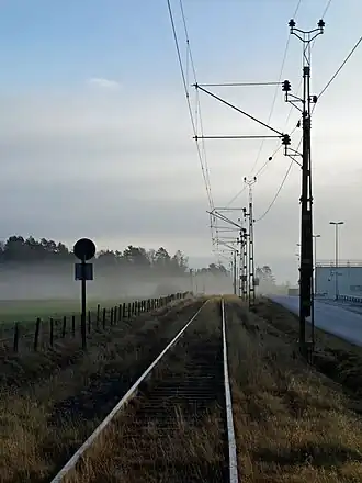 A railway line with overhead wires, supported by latticework gantries on its right, seen just from its side. On the left is the rear of a sign with a circular piece above an equally-sized rectangular one. Golden-colored grasses rise from in between the ties, and there is a fogged area of woodland around the tracks in the distance under clouds with blue sky above them. On the right is a road; a fence separates the track right-of-way from a green field on the left.