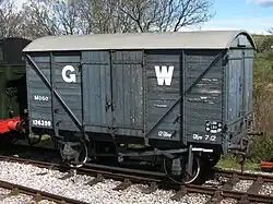 A short goods van built from planks of wood. The sides are mid-grey with a large white G to the left of the doors and a large white W to the right.
