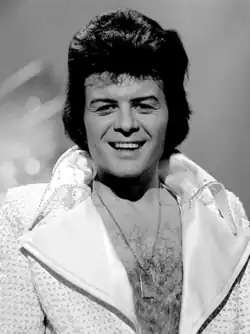 A black-haired man with a hairy chest, wearing a shiny jacket open to the waist, with large lapels, smiles towards the camera.