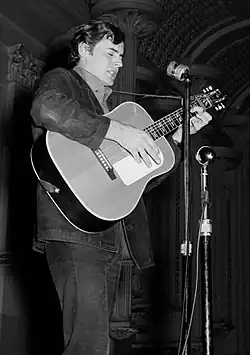 A man plays an acoustic guitar and sings into a microphone on a stand. He is shown in right profile, wearing dark clothes and looking downwards.