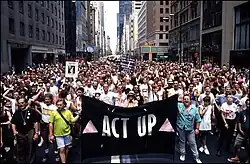 A large crowd of people is gathered behind a black banner that reads "ACT UP" between two pink triangles.