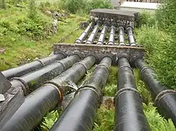 Looking down grassy hillside at six large black metal pipes, which pass through concrete blocks. The pipe sections are bolted together, with a small amount leaking out and spraying upwards at one joint
