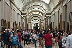 A high walled gallery with painting hanging from walls and a statue of Diana the Huntress is packed with visitors.