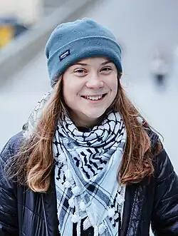 Portrait of Greta Thunberg. She is a young white woman with long blonde hair. She is wearing a black jacket, a knitted cap and a white keffiyeh around her neck.