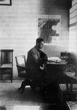 A black and white photo of Gu Jiegang seated at a desk in an office. A map of the area around Beijing is visible
