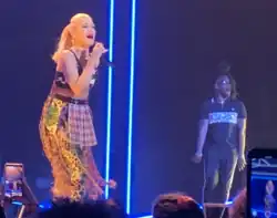 A color photograph of Gwen Stefani in a colorful outfit performing onstage with microphone in hand.