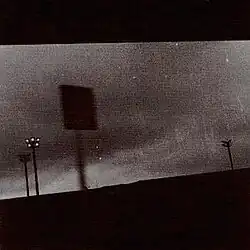 A grainy black-and-white photo of a sign in a parking lot with lights in the background.