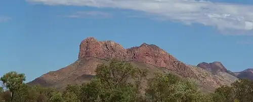 Daytime landscape photo, showing a range of hills with the nearest rising to a rocky red peak, below a blue sky with a few white strings of cloud, and above the tops of eucalyptus trees.