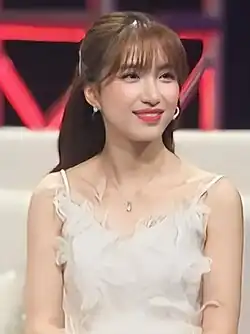 blurry image of Hòa Minzy wearing a white dress, sitting on what appears to be a couch onstage, grinning and looking left of camera