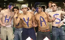 Five young shirtless men pose defiantly in a crowd. Each has a letter in blue on their chests to spell HOYAS.