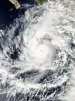 A photograph of a powerful hurricane off the Pacific coast of Mexico, approaching landfall. A thick rainband curves around the hurricane to its north and is beginning to spread over land, while a long arc of thin, high clouds is fanning out to the southeast, southwest, and northwest.