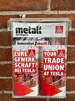 Close up of brick wall with attached plastic container. Inside are English/German language brochures. The IG Metall trade union brochures in German and English say "Eure Gewerkschaft Tesla bei Tesla" and "Your trade union at Tesla". Behind them is a larger colorful (mostly red) magazine titled "metall: Dein Magazin" with the IG Metall logo.