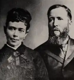 A portrait photograph of Ida Frances Hunt Udall (on the left) and David King Udall (on the right). Ida's hair is done up. She wears earrings, and the dress she wears features an ornate bow at the collar. David wears a necktie and suit, which is buttoned, and he has a long beard that reaches past his collar.