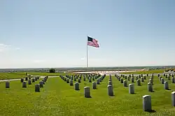 A United States flag flies behind a graveyard from a tall flagpole against a bright sky.