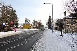 A ground-level photograph from a road with houses and trees visible, with a thin layer of snow covering the ground.