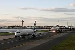 Five jumbo airplanes wait in a line on a runway next to a small body of water at John F. Kennedy Airport.