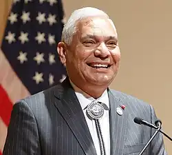 close-up of James R. Floyd wearing a gray striped jacket and white shirt with bolo tie, standing in front of microphone with US flag in background, grinning and looking left of camera