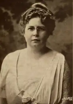 A white woman with light hair in a curly updo, wearing a light drapy dress and glasses