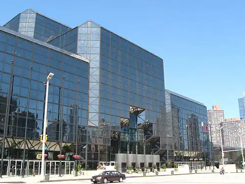 The Javits Center, a building of dark tinted glass, stands over a city street. The corners of the building are smoothed at 45-degree angles.