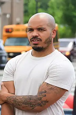 A young, black man with a mustache and anchor beard sits behind a laptop and stares off to his left. Another person, barely visible, is seated to Ellison's right.