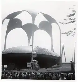 The theater as seen during the 1964 New York World's Fair. The theater itself has a large, disk-shaped roof. Around the theater, six tall metal columns hold up a canopy with a hole in the middle.