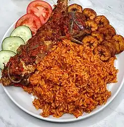 Jollof rice with fried fish and plantain, garnished with cucumber and tomatoes