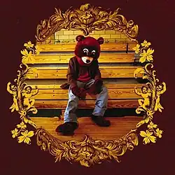 West wearing a bear mascot costume, slouched over on a bench. The image is surrounded by an intricate gold design, all against a brown background.