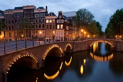 Photo of canal houses and two bridges with lights along their arches at a canal intersection at night