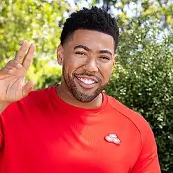 A bust-length photograph of a man in a red shirt waving with his right hand.