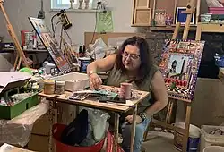 Kirsten Abrahamson in her studio, surrounded by art supplies, working on her piece: "June: Every Six Days (An Indigenous Women Goes Missing in Canada)". Meanwhile, as her daughter takes the candid photo: "July: The Red Balloon" sits on an art easel behind her. Photo taken: June 22, 2019 by her loving daughter.