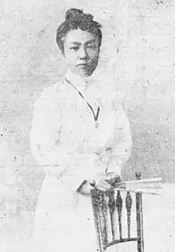 A young Japanese woman wearing a white gown, standing with a chair, holding a fan