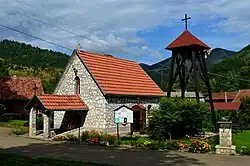 A picture of a small white stone church with an orange tile roof. The sun is high overhead and flowing plants are growing just in front of the church. A second building and green hills are visible in the background. A cross is above the church.