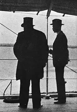 Kruger viewed in silhouette from behind, Bredell to his right. Kruger is wearing his top hat.