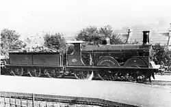 A black and white photograph of a locomotive and its coal-wagon facing rightwards at a station platform. On the wheel casing reads "JACOMB HOOD".