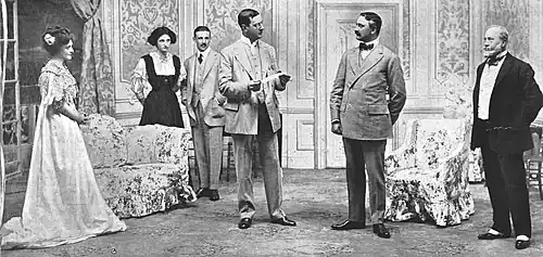 scene from a stage play with six characters – two women and three men – in Edwardian dress grouped in a luxurious drawing room