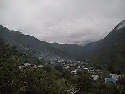 A photo overlooking Laprak, with many houses with various colored roofs, taken from a vegetated area of mountainside, with green mountains and a cloudy sky in the background
