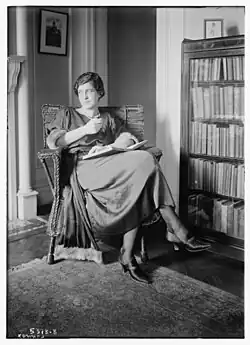 A white woman with dark hair, seated indoors, in front of a bookcase, with a book open in her lap