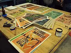 Colorful letterpress posters for "St Louis Microfest" spread out on a table alongside ink and other printing materials.