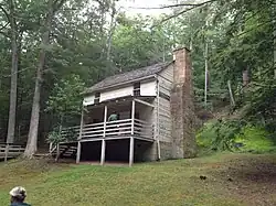 A two-story log cabin with a front porch and a forested hillside in the background.