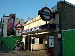 A building flanked by green, wooden walls with three people standing outside and one person walking by all under a clear, blue sky