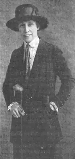 A smiling white woman wearing a black brimmed hat and jacket, with a collared shirt and necktie; she is standing and holding a golf club in her right hand