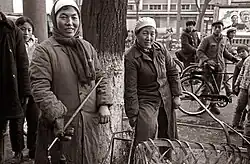 A black and white photo of two people in heavy coats, scarves, and hats holding tools smile candidly at the camera. Several others look at the camera from the background.